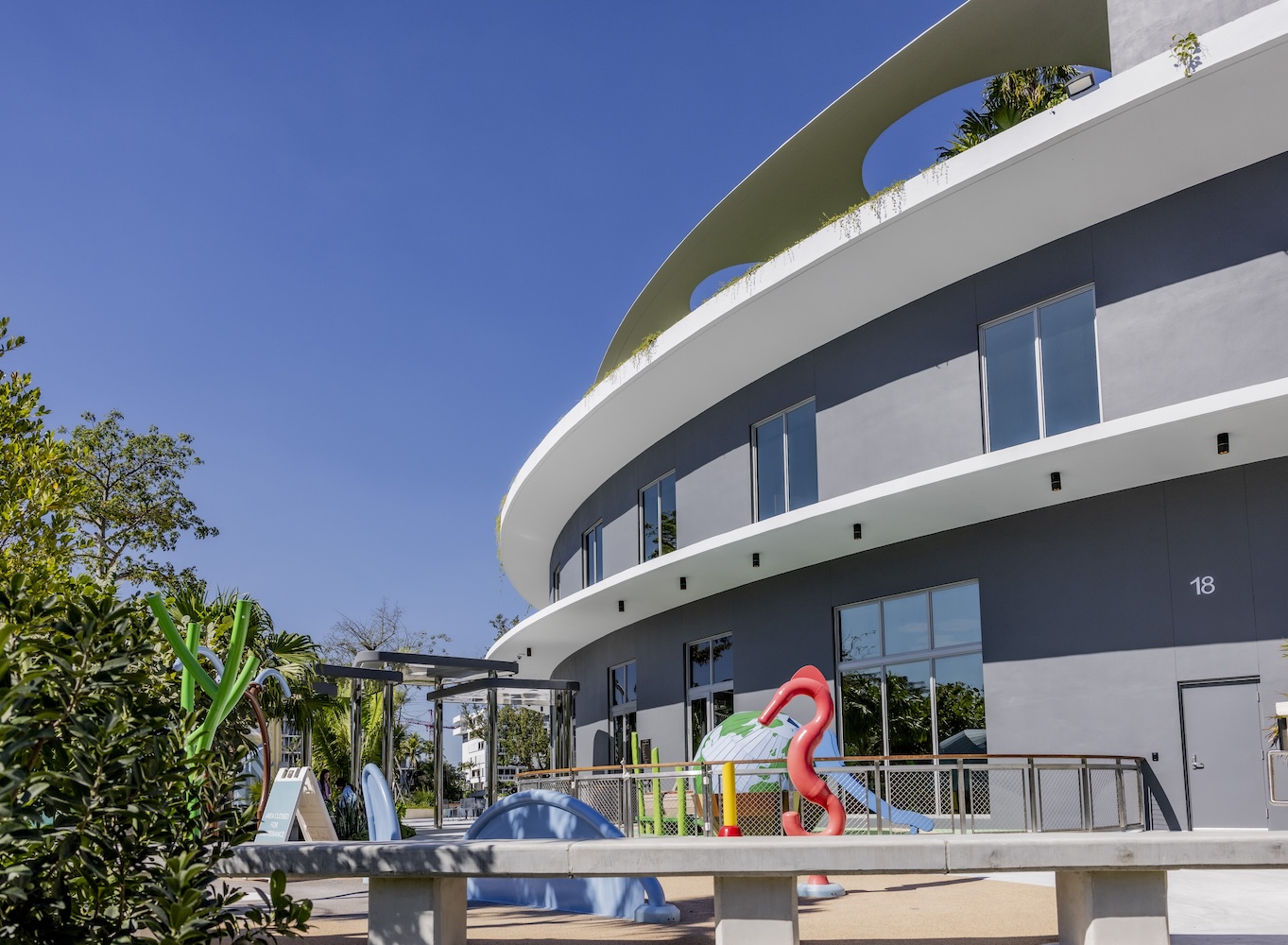 View of the Bal Harbour Waterfront Park. A winding bench, colorful water feature with brightly colored sea animals are visible in the foreground. In the background, the community center is seen along with lush landscaping, and a globe themed playground.