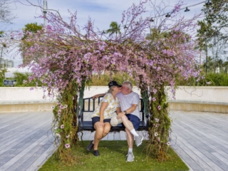 COMPRESSEDFleursDeVilles2024 PARK 03-02-24_61 (1) An older couple kissing on the bench with pink flowers all around them.
