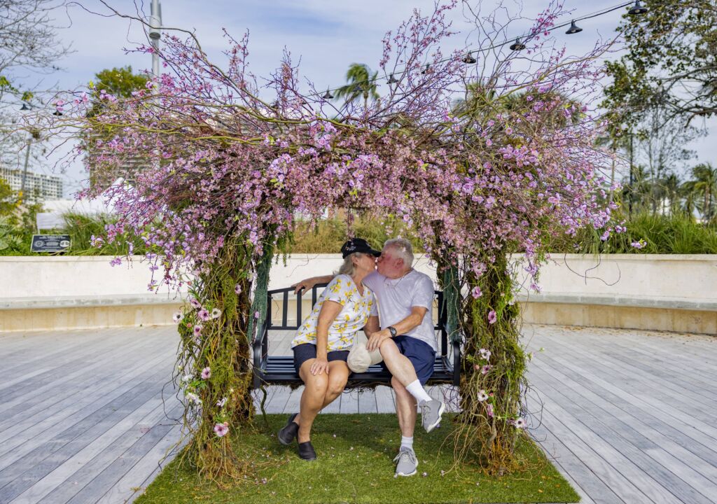An older couple kissing on the bench with pink flowers all around them.