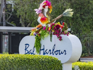 COMPRESSEDFleursDeVilles2024_12 (1) One of the Fleurs De Villes art pieces on top of a Bal Harbour sign. The art is of a woman sitting on a flower filled boat holding an oar.