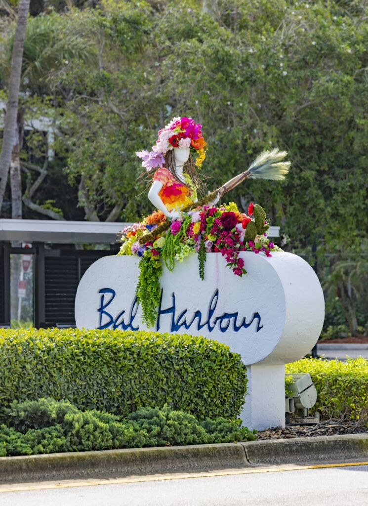 One of the Fleurs De Villes art pieces on top of a Bal Harbour sign. The art is of a woman sitting on a flower filled boat holding an oar.