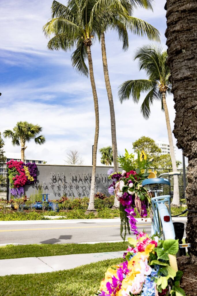 The back end of a blue bike with flowers all around it. The Bal Harbour sign is across the street in the background, aswell as palm trees.
