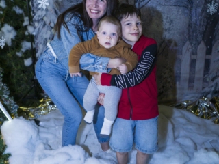 BalHarbourXmas-030-min A boy holding a baby, with his mother hugging both of them from behind. Standing in a winter photo op surrounded by snowflakes and a snow on the floor.