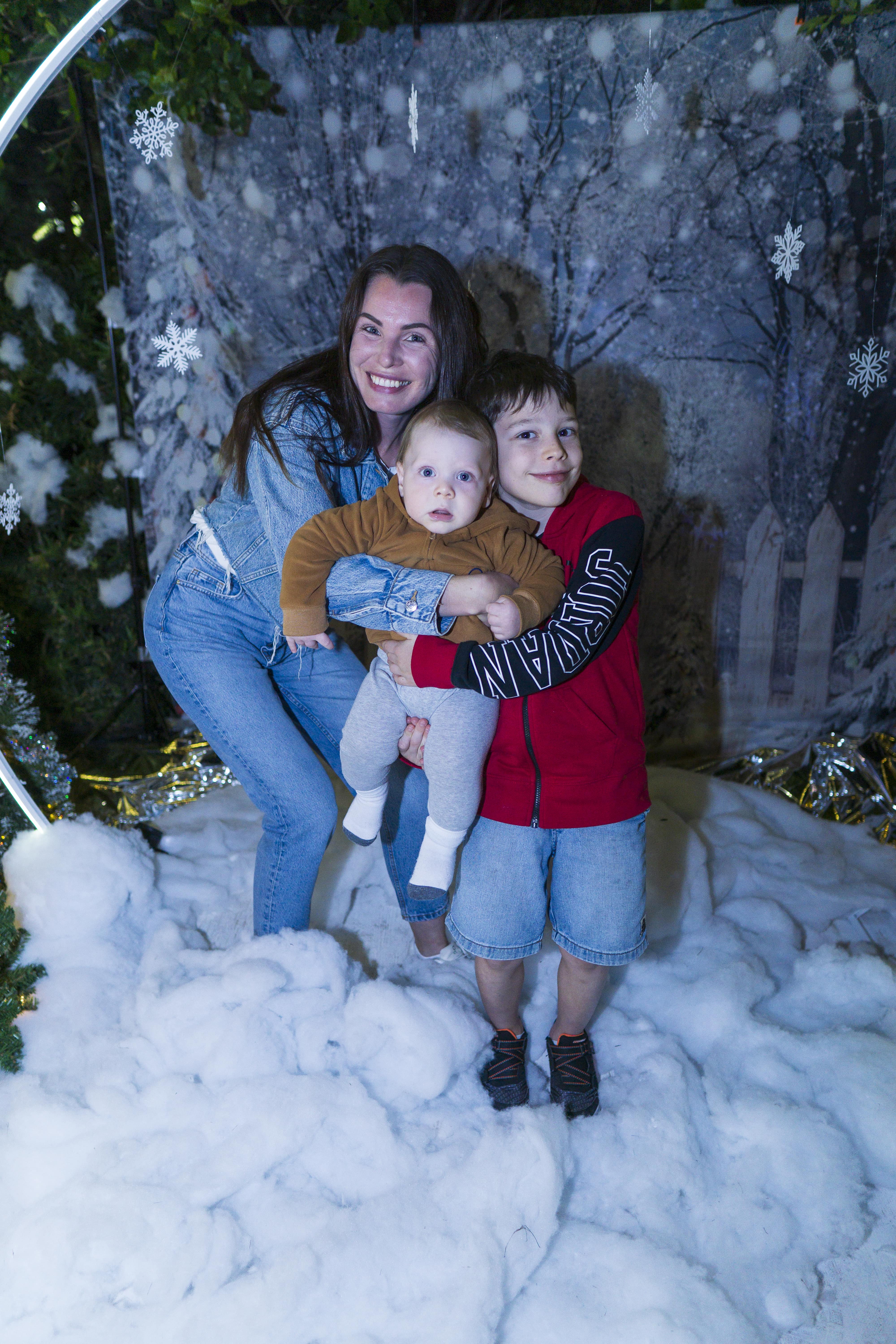 A boy holding a baby, with his mother hugging both of them from behind. Standing in a winter photo op surrounded by snowflakes and a snow on the floor.