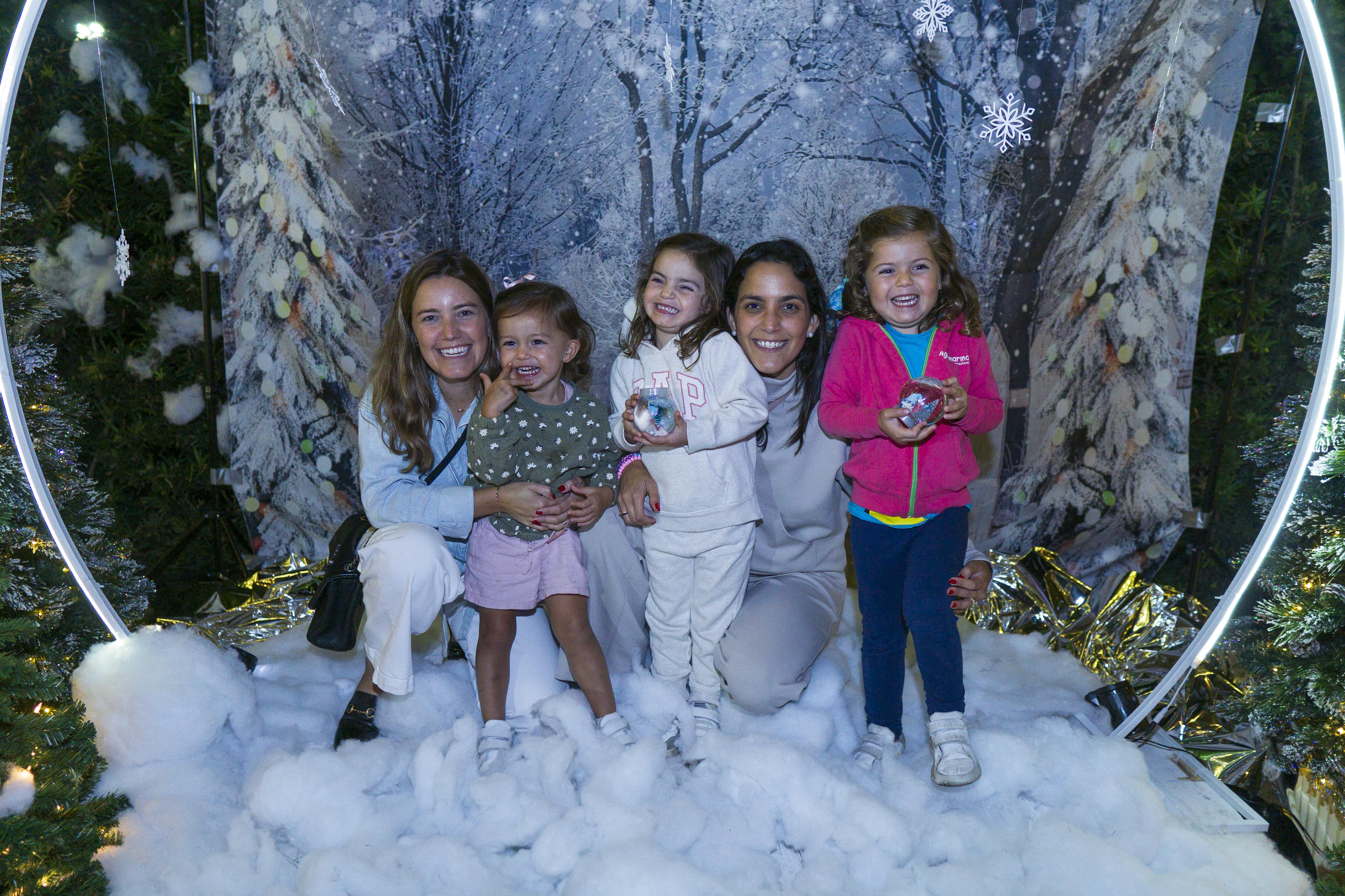 Two women crouched down on the floor hugging three little girls, all smiling with joy. Standing in a winter photo op surrounded by snowflakes and a snow on the floor.