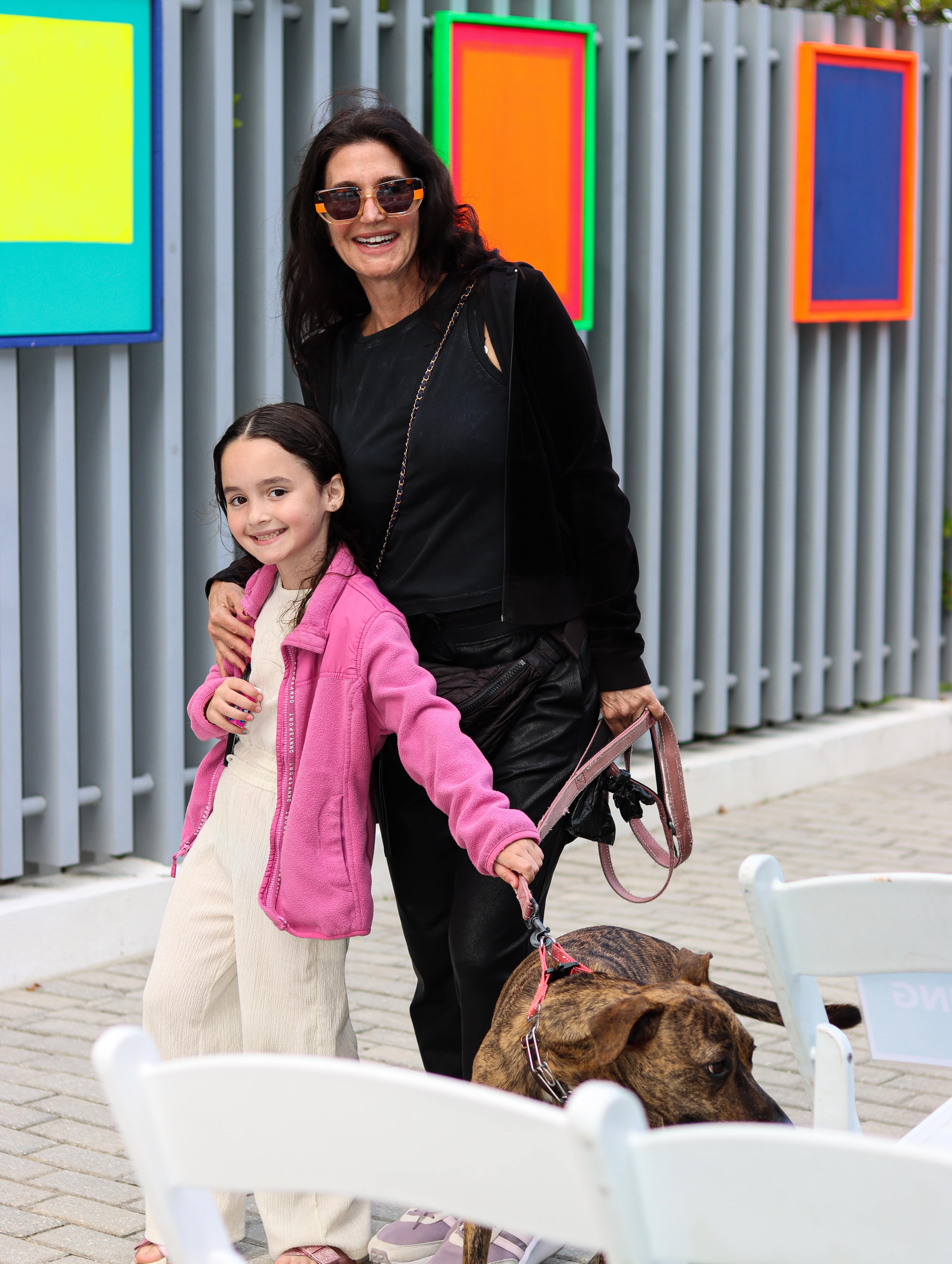 A mother holding onto her daughter, both grabbing the pink leash of a dog. Smiling for the camera in front of three paintings at the Dreaming In Color Exhibit.