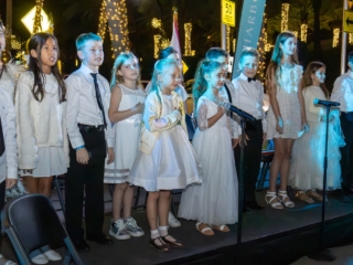 IMG_5939-Enhanced-NR-min A group of children standing on a stage, all dressed in white, singing holiday songs.