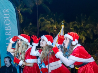 IMG_6002-Enhanced-NR-min Three girls all dressed in holiday outfits, with Santa hats and red dresses. They are singing to the crowd, mics in hand, all holding their other hand over their foreheads as if they are looking for something.