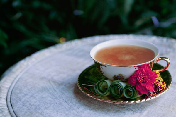 A delicate teacup filled with steaming tea, resting on a small wooden table surrounded by lush palm leaves. The vibrant green foliage creates a tropical, serene atmosphere, with sunlight filtering through the leaves, casting soft shadows over the scene, perfect for an outdoor tea experience.