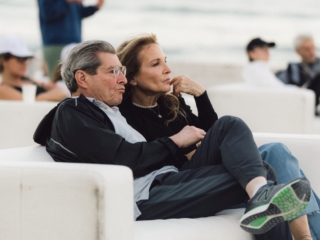 BalHarbourFilmFest-20-COMPRESSED A man and woman staring forward, and both crossing their legs seated next to each other on a white couch. You can see other couches with people in the blurred distance, as well as the ocean water and sand behind them.
