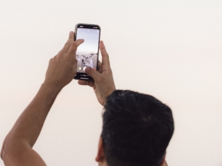 BalHarbourFilmFest-48-COMPRESSED The head and arms of a man holding up his phone, taking a photo. In the phone you can see couches and chairs lined up facing the movie screen. All around him all you can see is white.