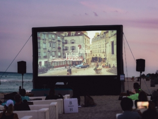 BalHarbourFilmFest-53-COMPRESSED1 The large screen projecting the movie is playing the title screen where it says "Julie and Julia" with a street and buildings shown. You can see a pink sunset behind the screen and white couches with residents seated beneath it.