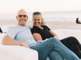 BalHarbourFilmFest-7-COMPRESSED A man and woman sat next to each other, smiling at the camera. They are sitting on a white couch and you can see the sand and water behind them.