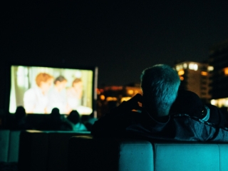 BalHarbourFilmFest-73-COMPRESSED A man and woman are seated next to each other on a couch, with another couch farther in front, both illuminated by a blue glow. You can only see the back of their heads with the only other lighting being the movie screen and buildings in the distance.