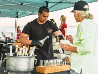 BalHarbourFilmFest-9-COMPRESSED2 A man working under a teal tent holding a champagne bottle in one hand and offering a glass of champagne in the other hand to a woman. You can see other residents behind the tent conversing and the blue ocean water.