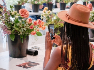 flower frame workshop-COMPRESSED The back profile of a woman wearing a coral-colored hat taking a photo of her beautiful floral arrangement in a large black vase. You can see other floral arrangements lined up on a table in the blurred distance.