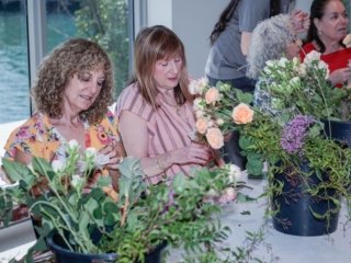 flower frame workshop-COMPRESSED3 Women work diligently next to each other on their floral arrangements. You can see the ocean water through the window behind them.