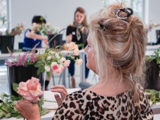flower frame workshop-COMPRESSED6 The side profile of a woman with her hair held up, she is holding a flower in one hand and staring down at it. You can see her floral arrangement placed on the table in front of her, with other residents working on their own creations in the background.