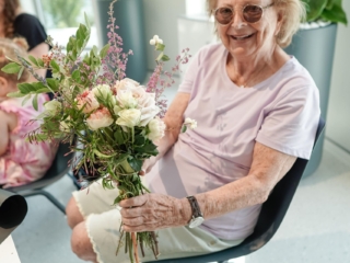 flower frame workshop-COMPRESSED7 A woman is seated and holding her floral bouquet in her hands. She is wearing a light pink shirt and glasses, smiling at the camera.