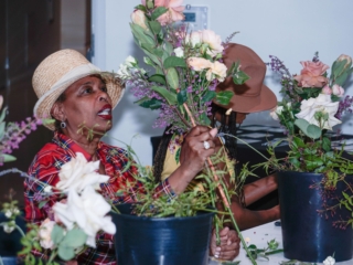 flower frame workshop-COMPRESSED8 A woman is staring intently at her floral arrangement, holding it in her hand. Another woman is working beside her, both wearing hats, and you can see their flower buckets placed on the table.