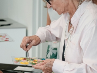 flower frame workshop-COMPRESSED9 A focused woman placing a flower with a small tool on the clear floral frame she is holding. You can see other yellow and white pressed flowers already on her creation, as well as different leaves and frames on a black table under it.