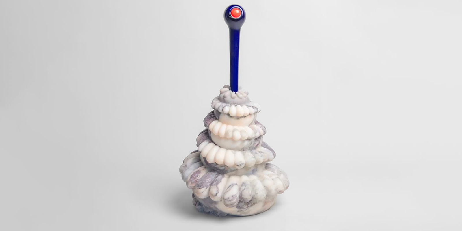A colorful, layered sculpture resembling a twisted dessert, topped with a blue handle and red knob, set against a plain white background.