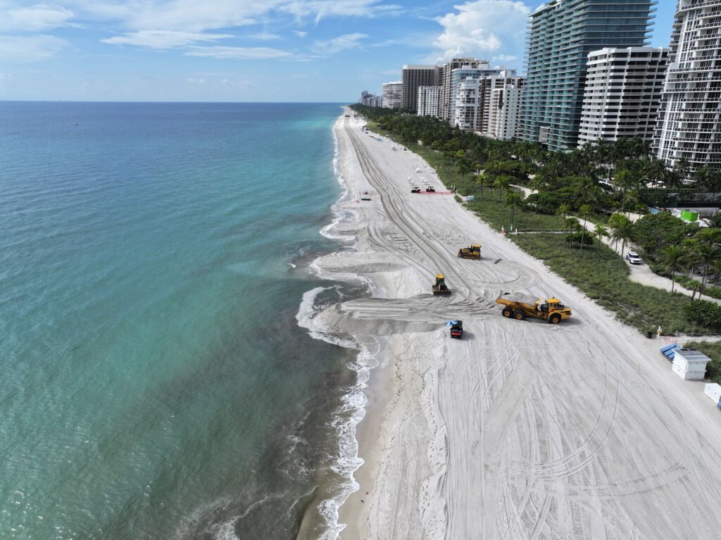 Aerial view of Bal Harbour Beach in Miami with turquoise ocean water, sandy shoreline, and construction equipment working on beach renourishment near oceanfront condominiums and palm trees.