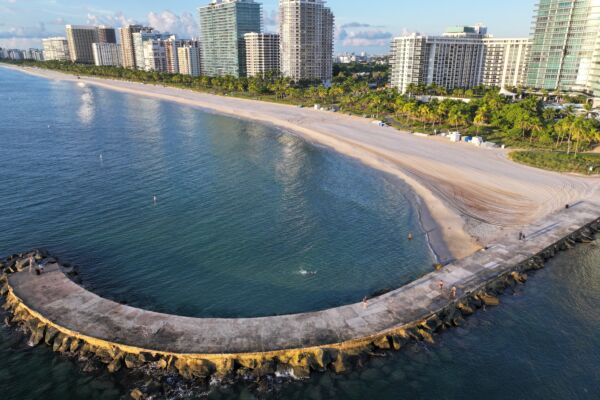 Aerial view of a calm beach with gently lapping waves curving along high-rise buildings. A curved pier juts into the sea, under a clear blue sky.