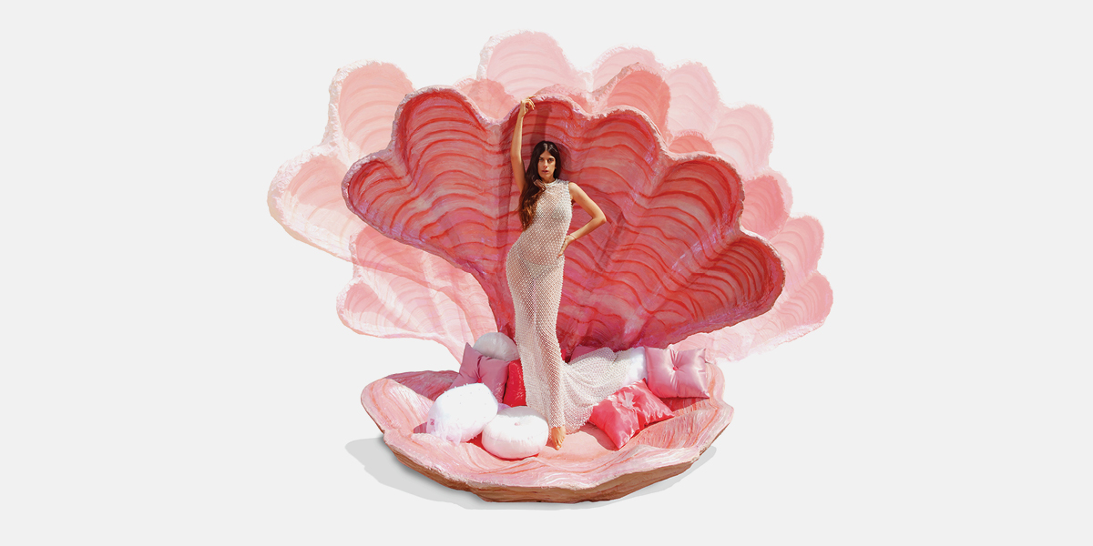 A woman in a form-fitting dress poses elegantly inside a giant open clamshell with pink and white hues. The scene conveys glamour and whimsy.