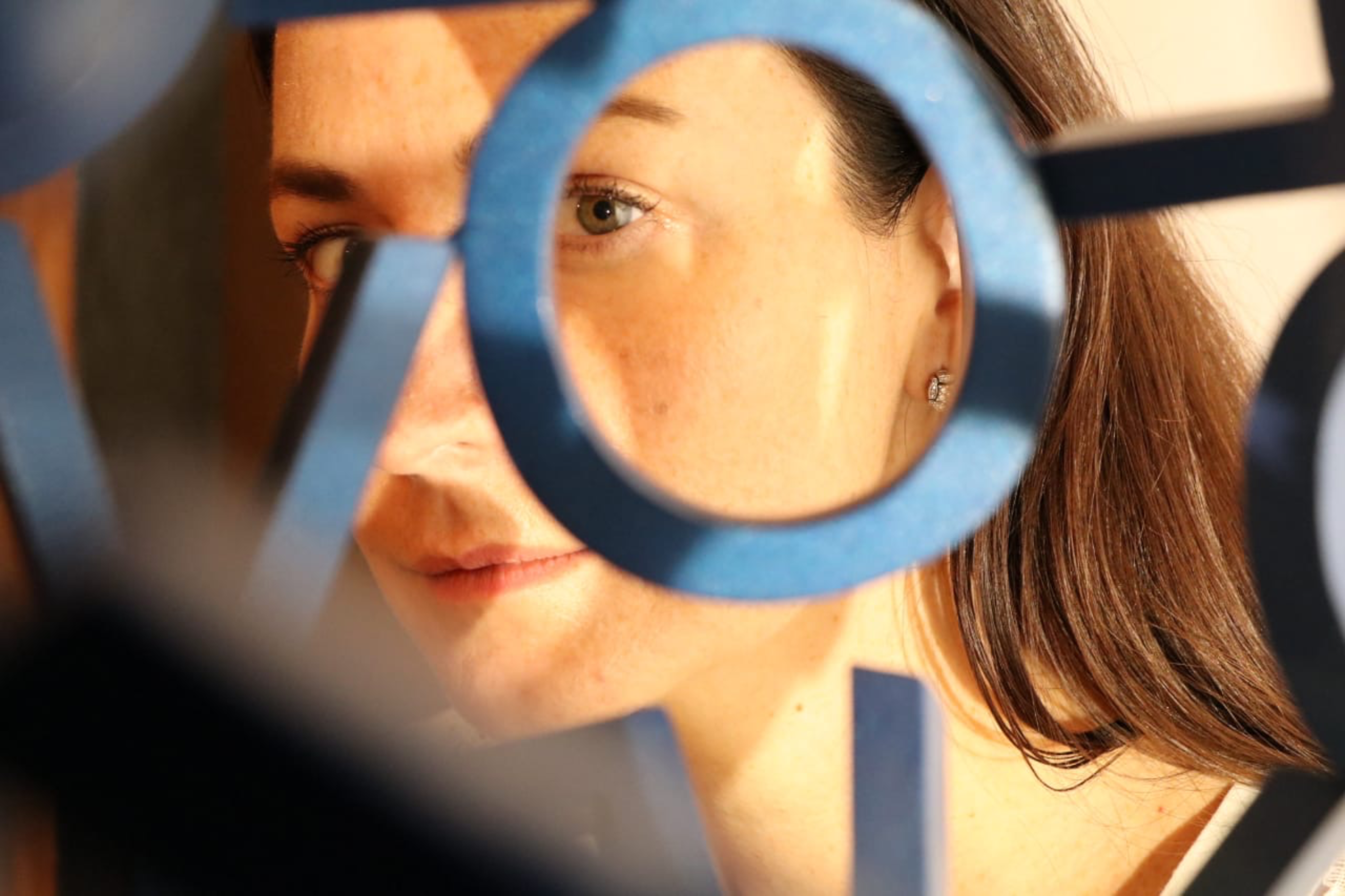 A woman's face is partially visible through a geometric blue sculpture. Soft lighting highlights her relaxed expression and natural complexion.