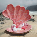 eight foot tall oyster shell open in shades of pink with pillows inside sitting on a beach