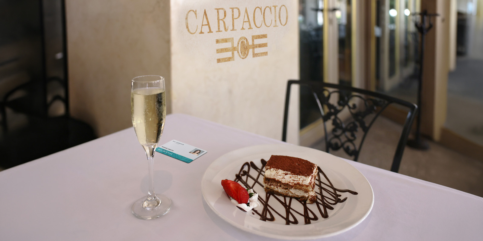 A table at Carpaccio restaurant features a glass of champagne and a chocolate-drizzled tiramisu slice with a strawberry on a white plate. Elegant ambiance.