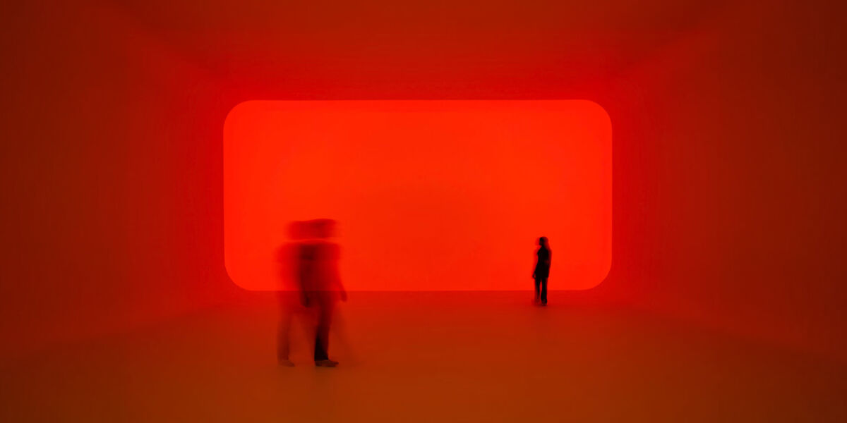 A red-lit immersive art installation featuring a large glowing rectangular light panel. Two silhouetted visitors stand in the space, with one figure blurred in motion in the foreground, creating a dramatic, atmospheric effect.