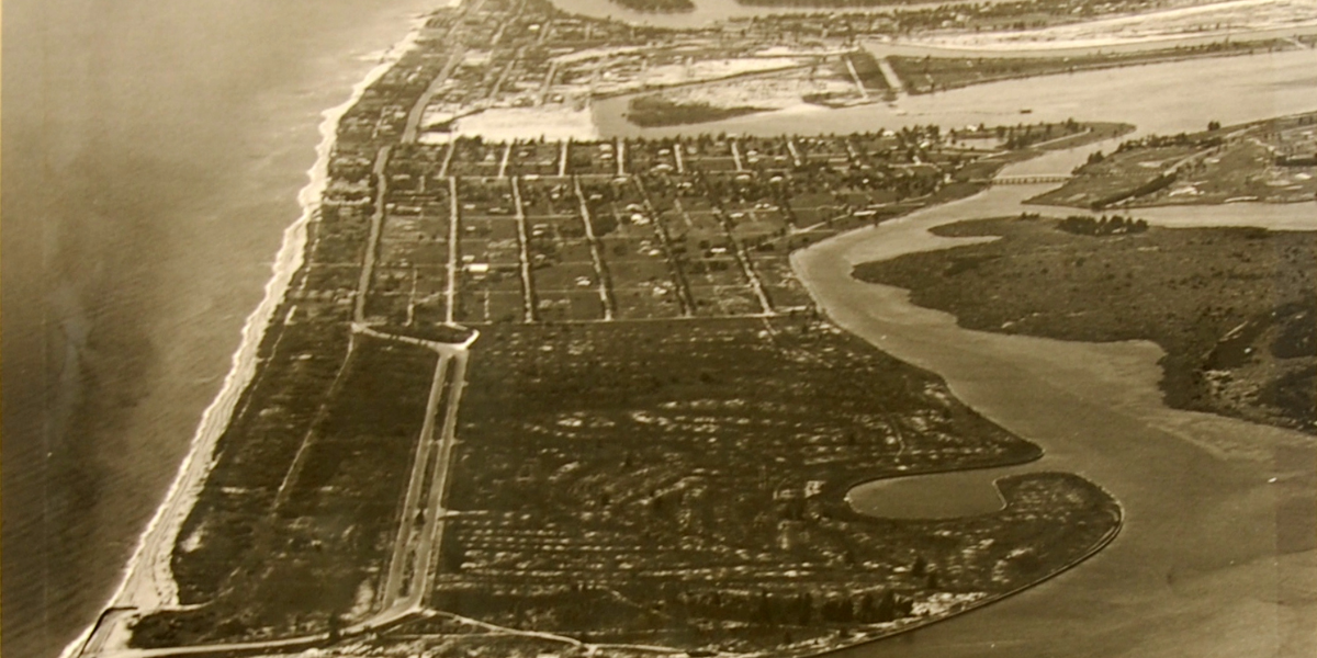 Sepia-toned historic aerial photograph of a coastal community, showing a long stretch of sandy beach, a grid of streets and residential lots, undeveloped land, and winding waterways along the shoreline.